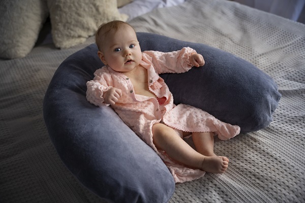 bebelus imbracat in roz, sprijinindu-se de o pernuta gri, pusa pe un pat