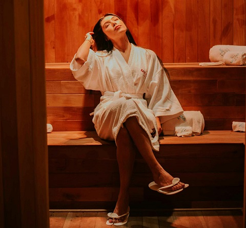 femeie tanara in halat de baie alb si papuci relaxandu-se intr-o sauna uscata