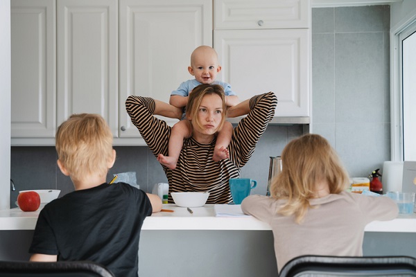 mama care isi tine bebelusul pe umeri in timp ce isi supravegheaza copiii mai mari care isi face temele in bucatarie