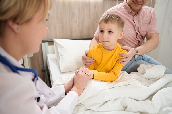 doctorita care ia de manuta un baietel trist care se afla pe un pat de spital alaturi de un barbat