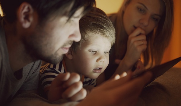 parinti si baietel care se uita pe tableta in pat, pe intuneric