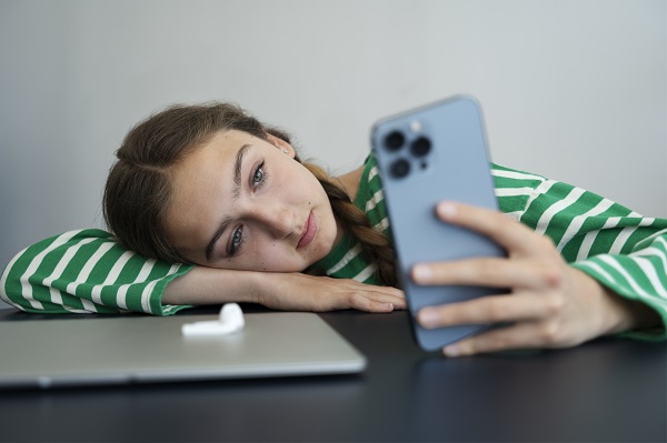 adolescenta deprimata stand cu capul pe birou si uitandu-se pe telefon