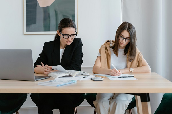 doua femei tinere cu ochelari studiind si luandu-si notite stand la birou