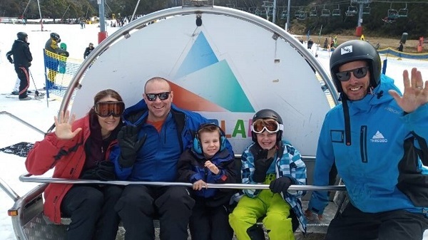 familie imbracata in echipament sportiv de iarna aflata pe pista cu zapada in jur fac cu mana la camera