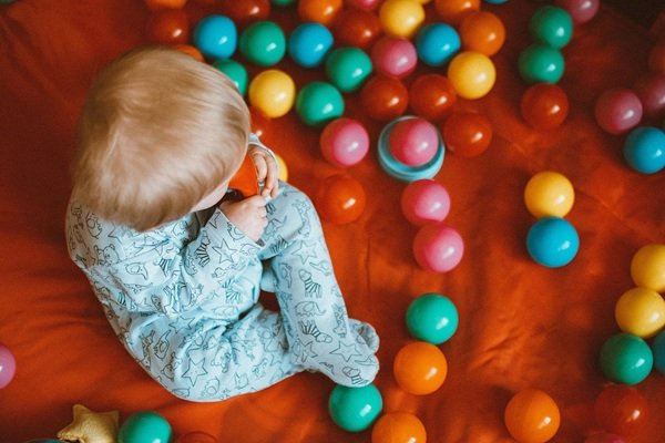 copil mic imbracat in albastru se joaca oe covor cu bile colorate de plastic