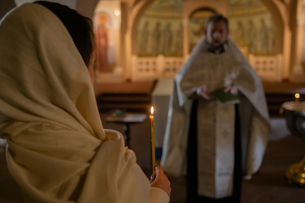 o femeie cu batic pe cap tine lumanare in mana in timp ce preotul se roaga in biserica